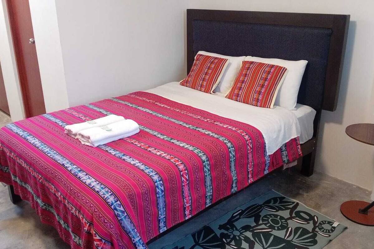 A bed with a patterned bedspread and pillows, a bedside table with a lamp, and a rug on the floor.