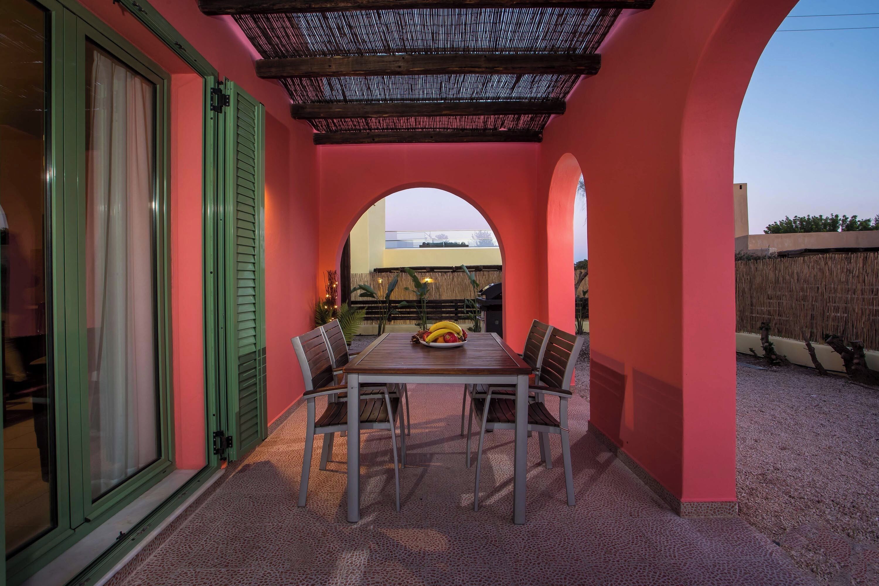 A covered patio with a table and chairs, an archway, and a view of a building in the background.