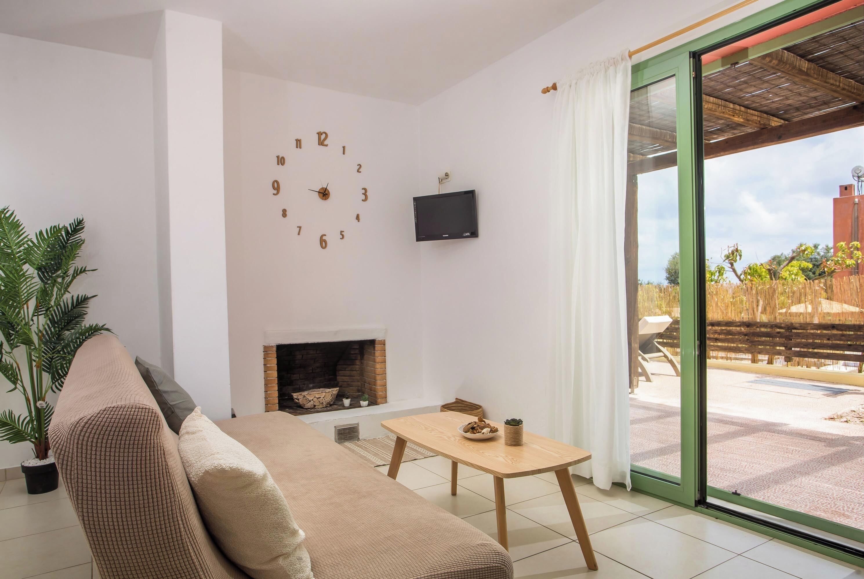Uma sala de estar com um sofá, uma mesa com uma tigela e uma planta, uma lareira, um relógio e uma TV na parede.