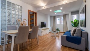 Una sala moderna con piso de madera, un comedor con mesa y sillas, un sofá, un televisor y un ventanal con cortinas.