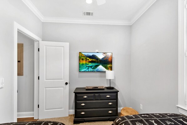 Une chambre avec une porte blanche, une commode noire dotée de tiroirs, un téléviseur fixé au mur qui diffuse un paysage pittoresque, et une lampe posée sur la commode.
