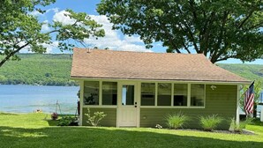 A small, single-story cabin with a sloped roof, large windows, and a door. It is surrounded by a well-maintained lawn and a body of water in the background.