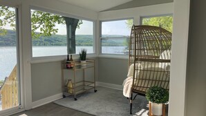 A room with large windows overlooking a lake, a wicker chair, a small shelf with decorative items, and a white rug.