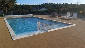 A rectangular swimming pool with a metal safety railing, surrounded by a textured gravel area. There are lounge chairs and a covered seating area in the background. - Chalet for 4, terrace & heated pool (Pont-l´Abbé)