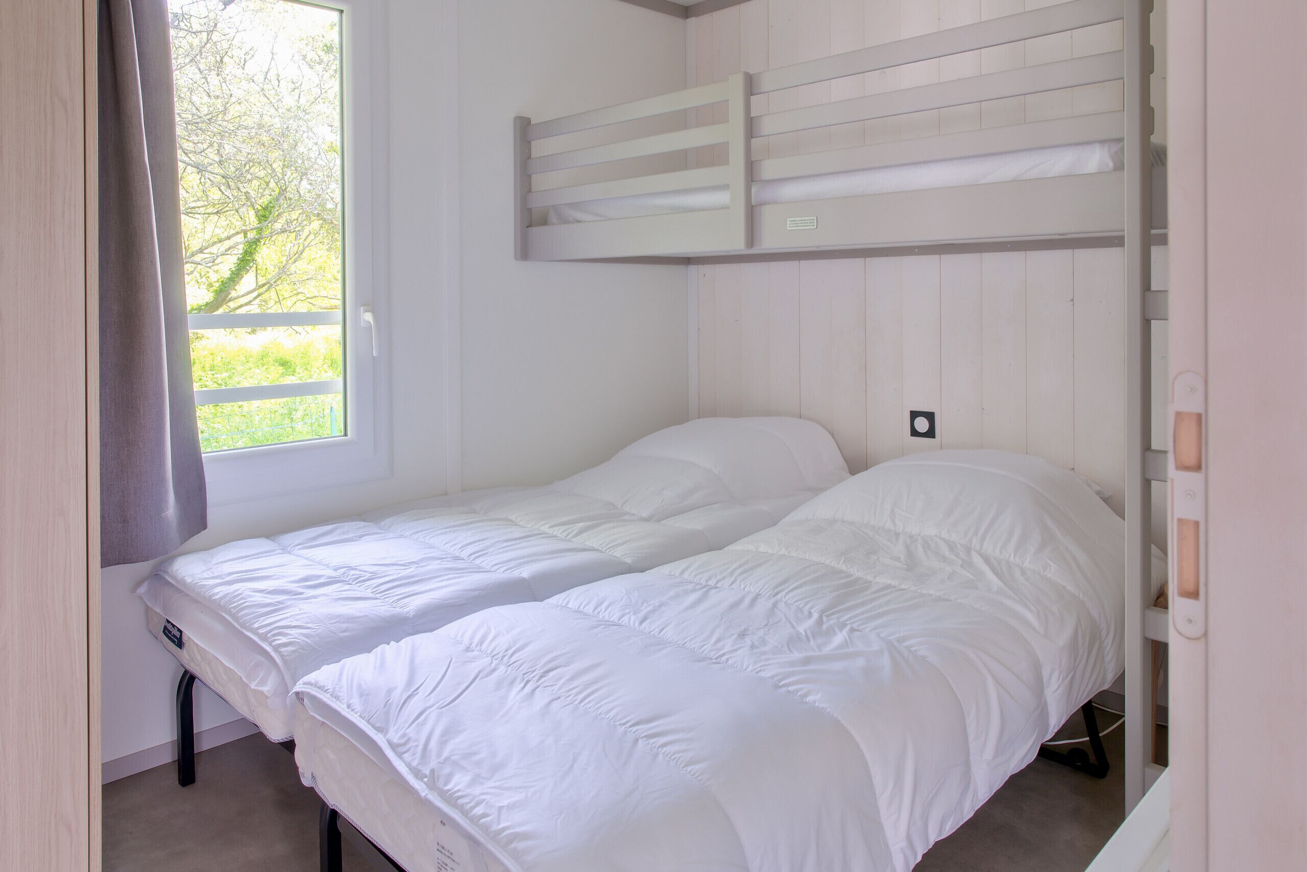Une chambre avec deux lits simples, une fenêtre donnant sur un espace verdoyant et un banc intégré au-dessus des lits.