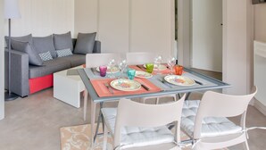 A dining area with a glass table set for two, a grey sofa with cushions, and a white floor lamp. - Chalet for 4, terrace & heated pool (Pont-l´Abbé)