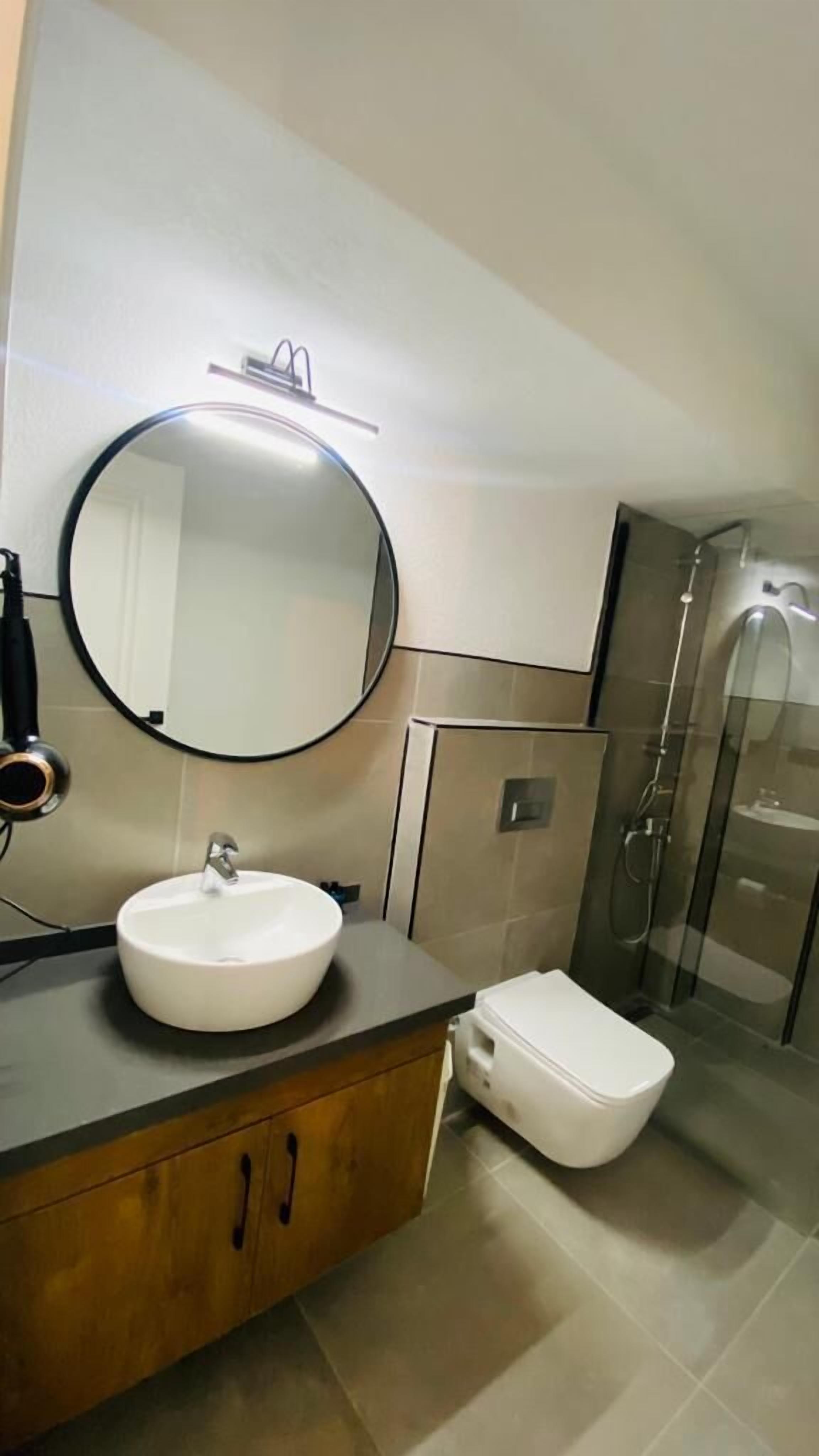 Een moderne badkamer met een ronde spiegel, een witte wastafel, een toilet en een douchecabine van glas.