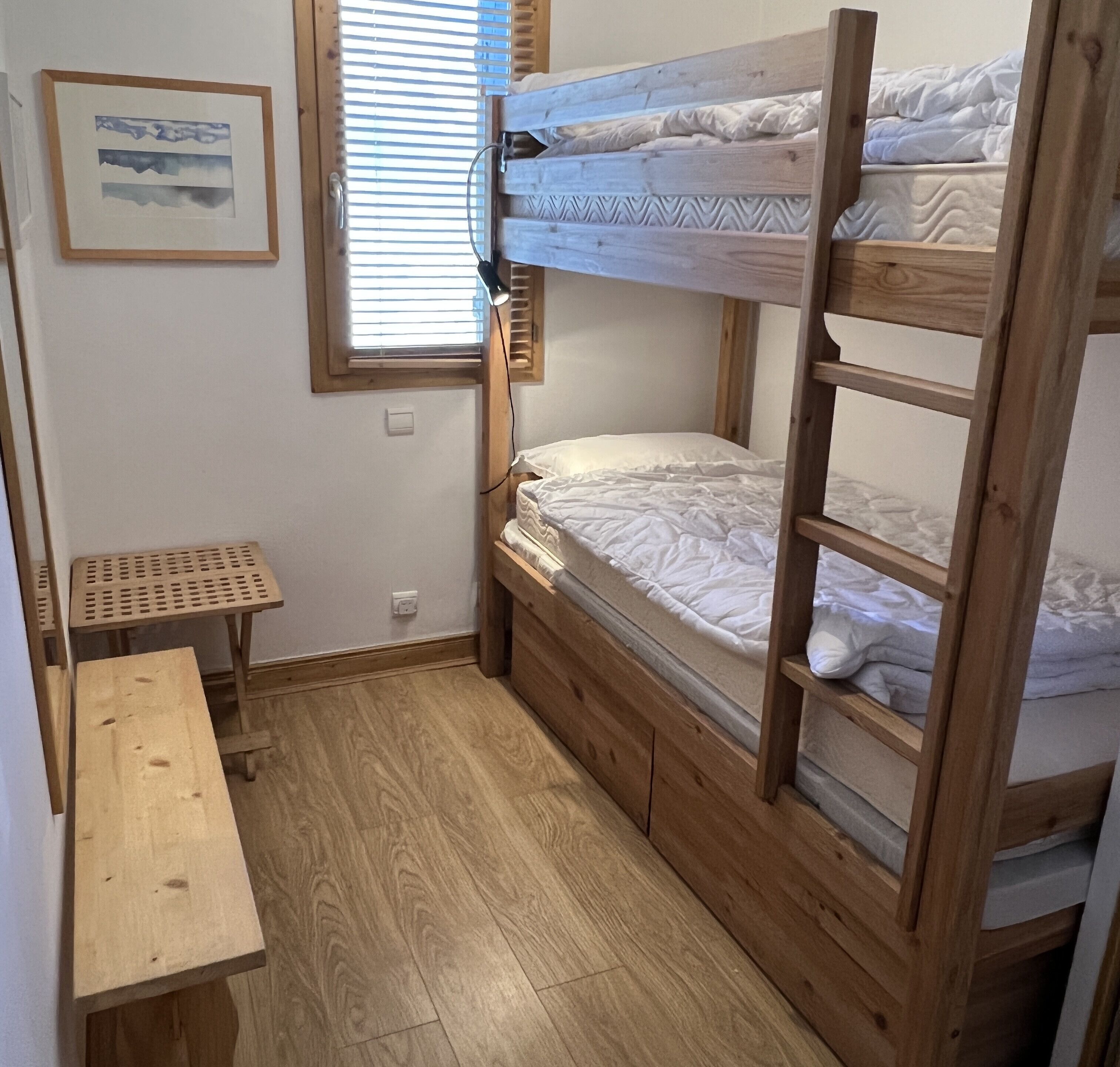 A bunk bed room with a wooden floor, a small wooden table, and a window with blinds.