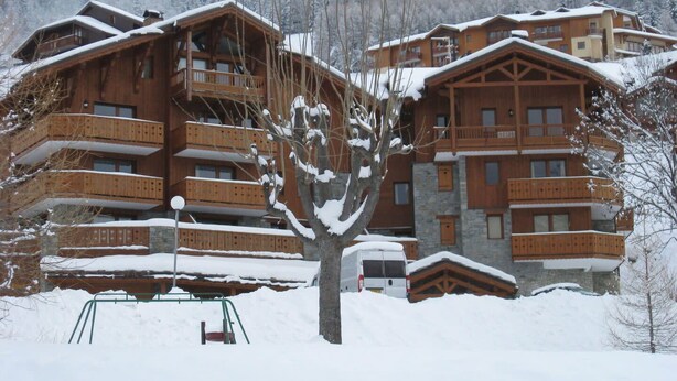 A wooden chalet-style building with multiple balconies, surrounded by snow-covered trees and a forested backdrop.