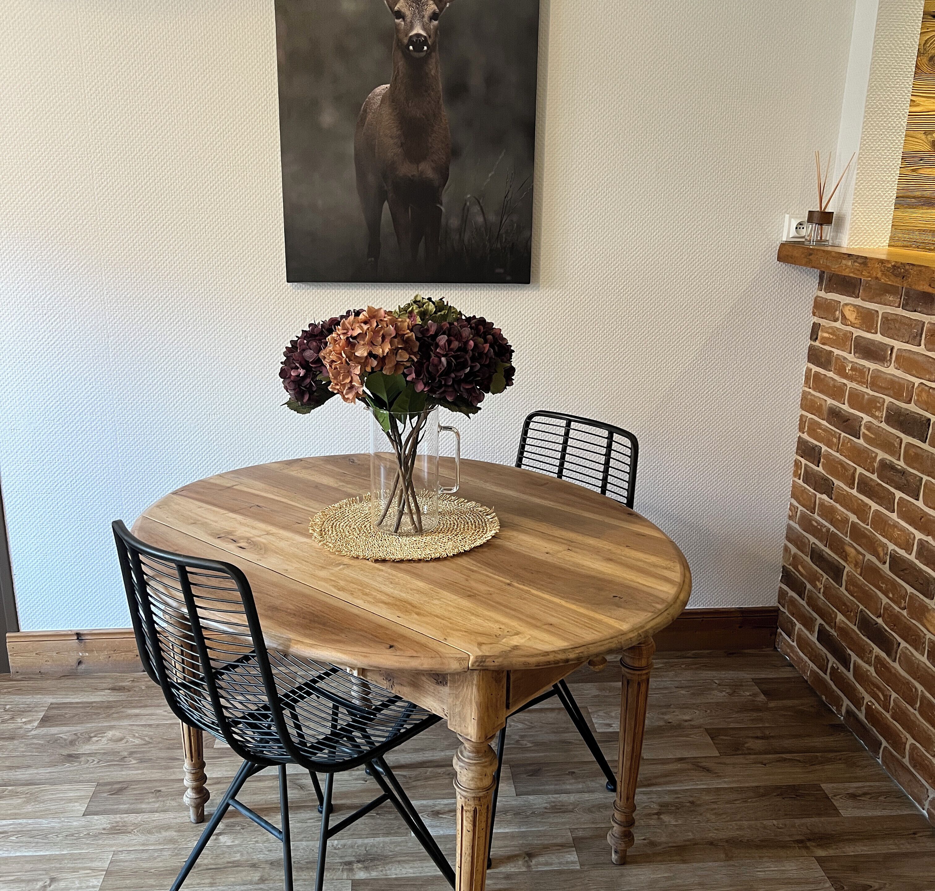 A round wooden dining table with a woven placemat, surrounded by black mesh chairs. A vase with flowers is placed on the table.