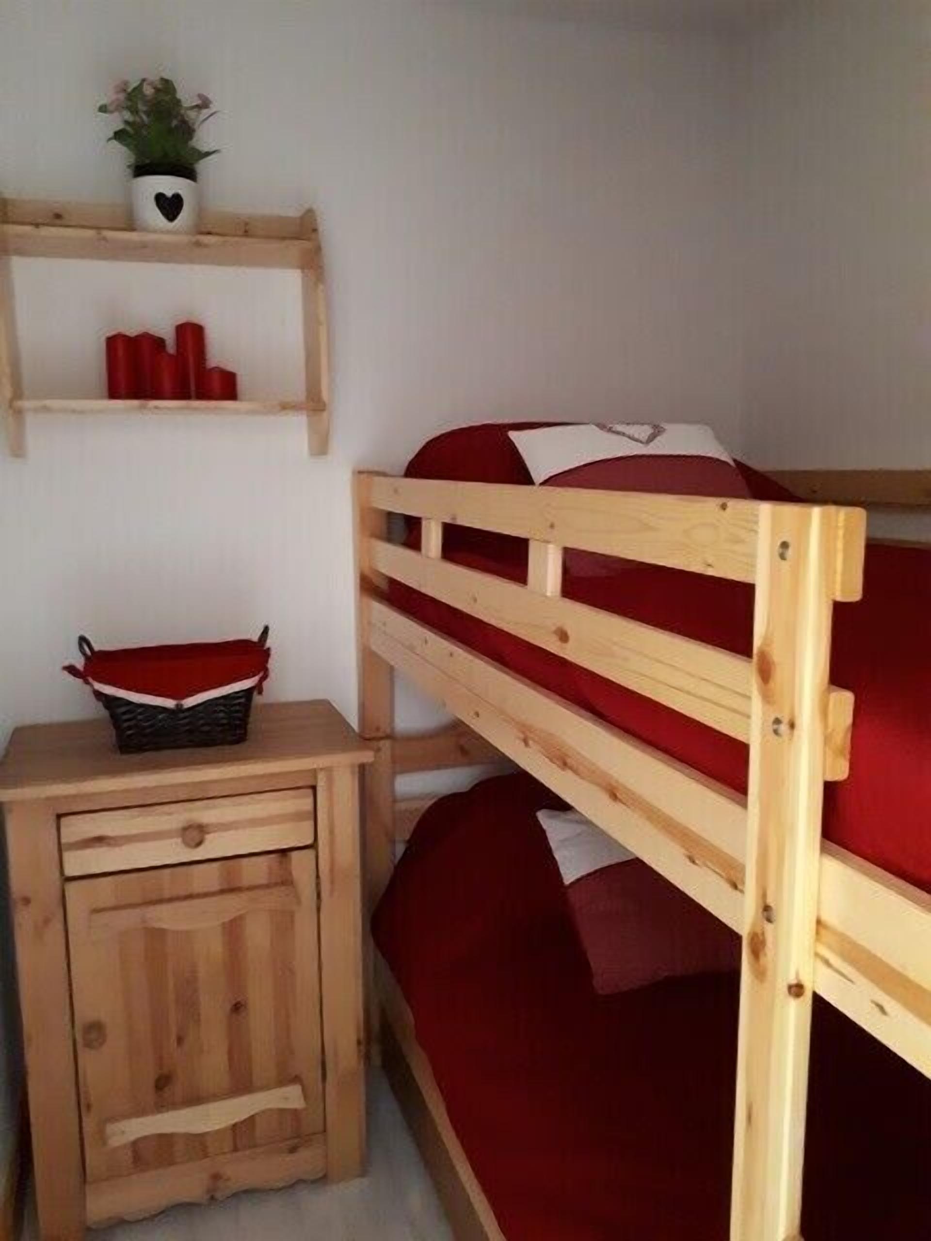 Una cama litera con colchón rojo, una mesita de noche de madera con cajón, y una repisa con planta en maceta y velas rojas.
