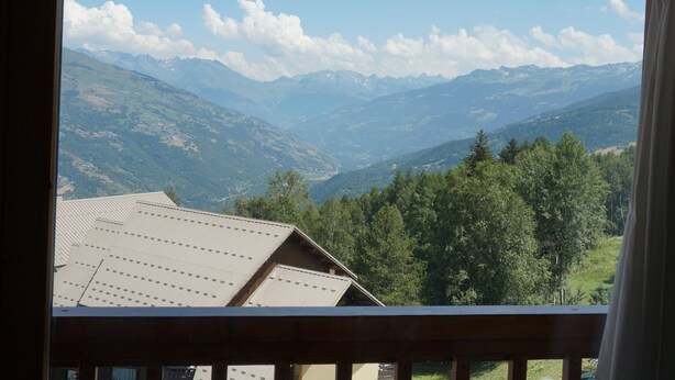 Vista de varanda com paisagem montanhosa, incluindo uma casa com telhado inclinado e guarda-corpo de madeira.
