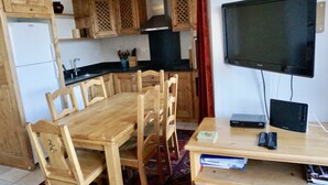 Uma cozinha compacta com armários de madeira, uma mesa de jantar com cadeiras, geladeira, fogão e uma TV de tela plana em um suporte.