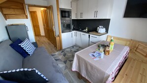 Ein kompakter Wohnbereich mit Küchenzeile, einer Couch mit gemusterten Kissen, einem Esstisch mit Tischdecke und einem an der Wand montierten Fernseher.