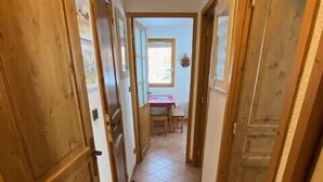 Ein schmaler Korridor mit Holztüren und Fliesenboden, der zu einem Raum mit Fenster und Tisch führt.