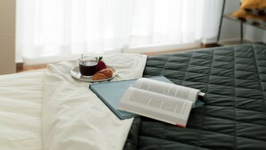 Una cama con un edredón verde oscuro, un libro, un vaso de líquido oscuro y un pastel en una bandeja.