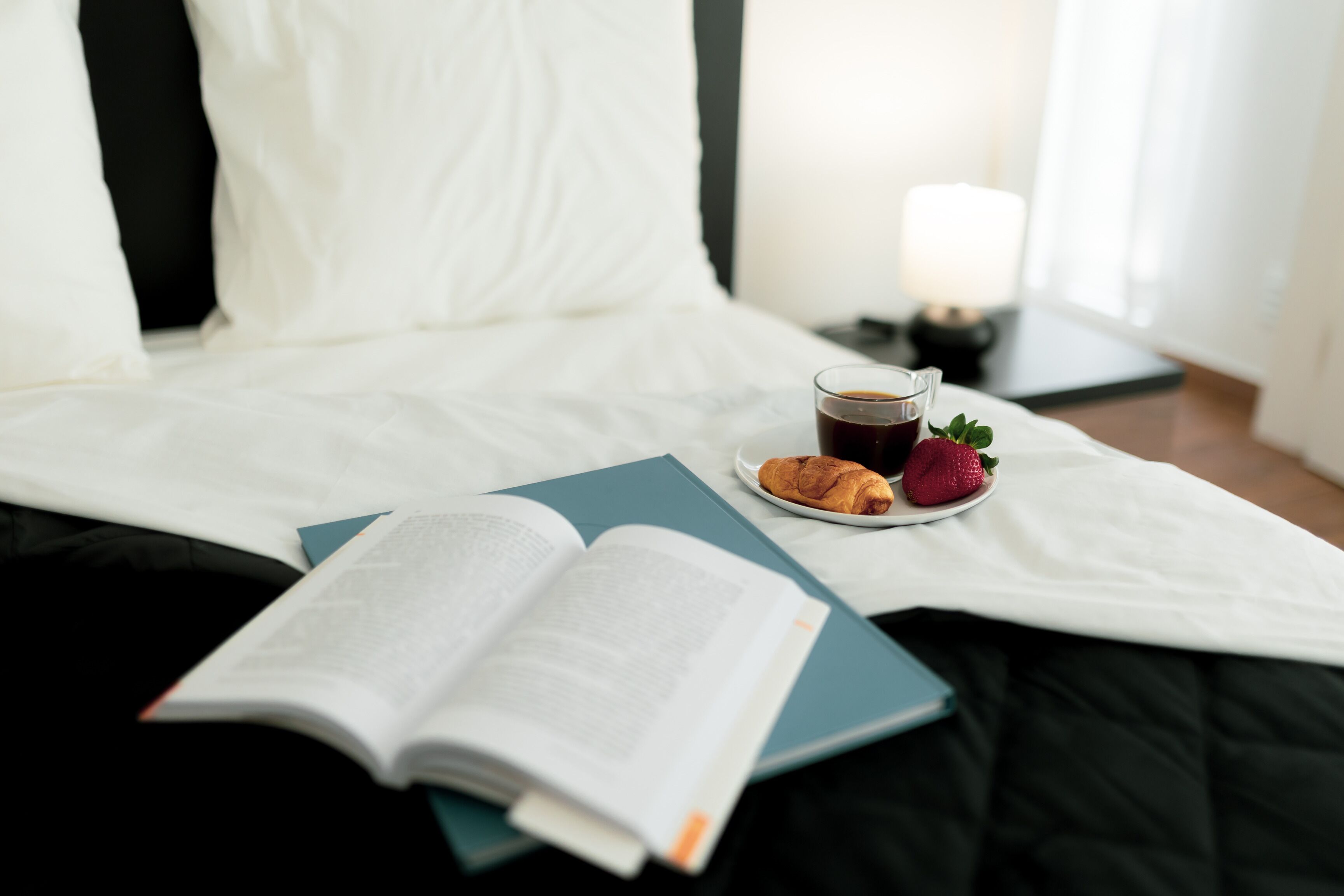 A neatly made bed with a book, a plate with a croissant and a strawberry, and a glass of coffee.