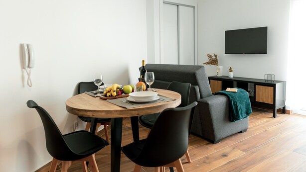 Una sala moderna con una mesa de comedor, sillas negras, un sofá gris y un televisor de pantalla plana montado en la pared.