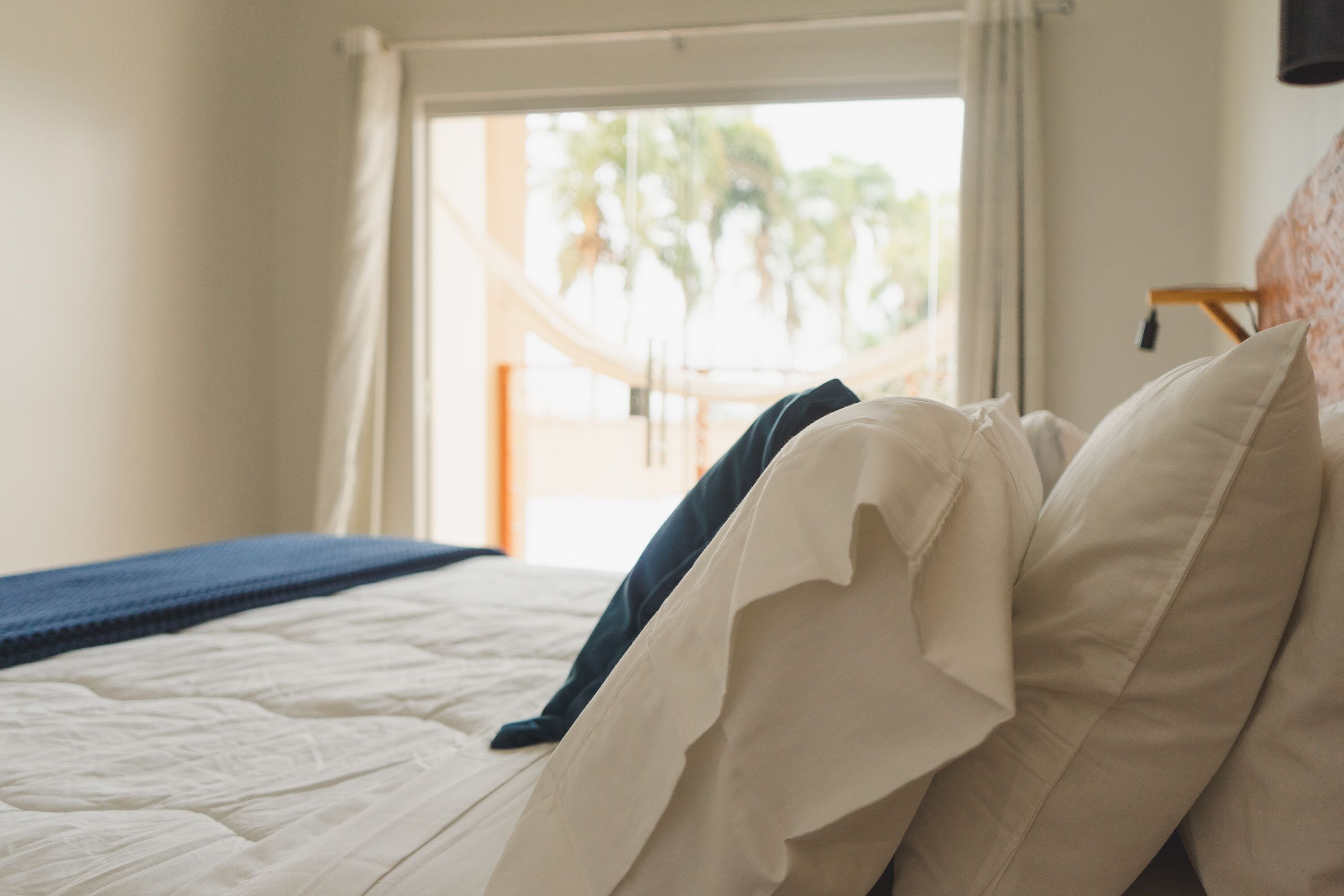 Una cama con ropa de cama blanca y una manta azul, una ventana con cortinas y vistas a palmeras afuera.