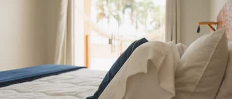 Una cama con ropa de cama blanca y una manta azul, una ventana con cortinas y vistas a palmeras afuera.