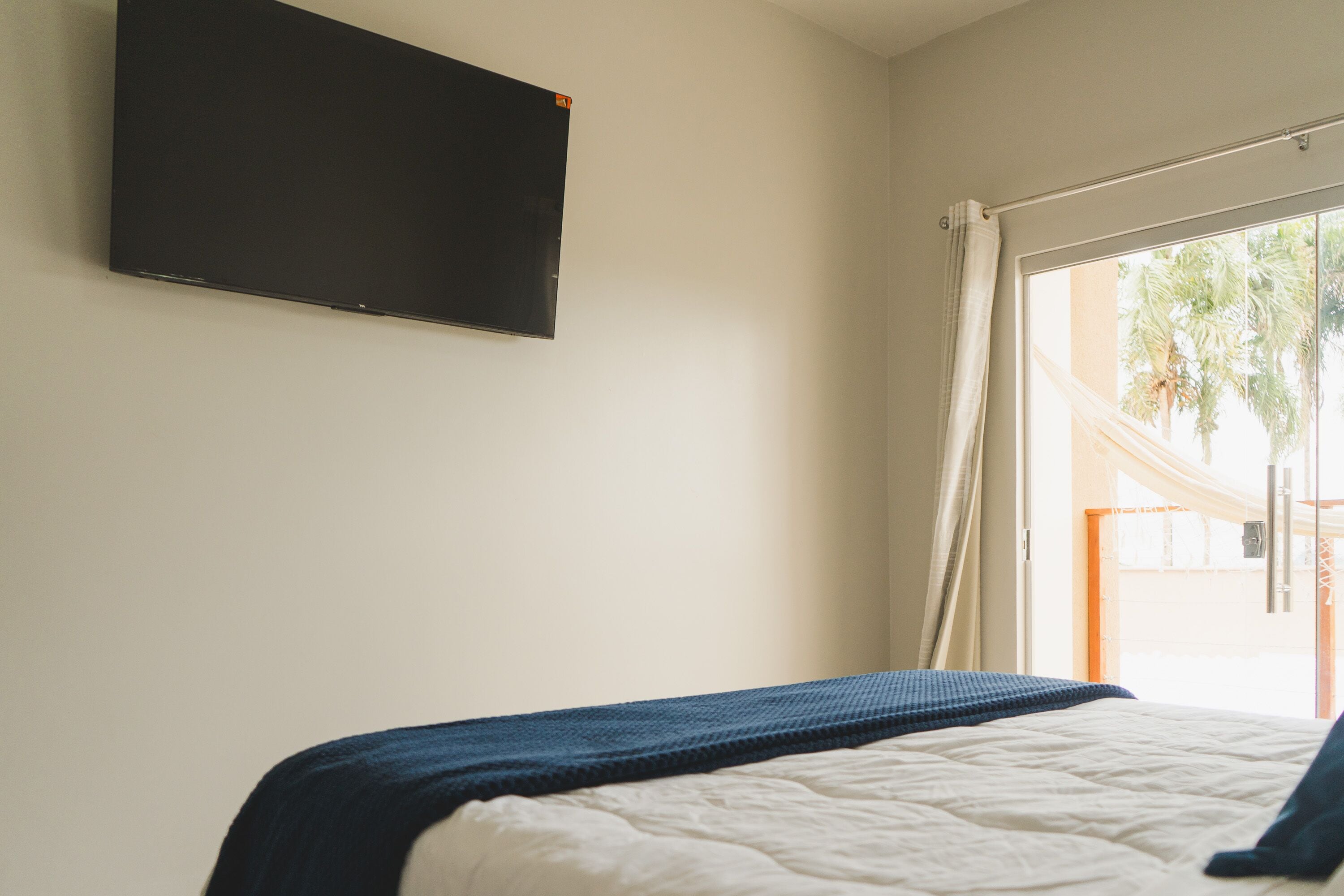 A bedroom with a bed, a flat-screen TV mounted on the wall, and a window with curtains.