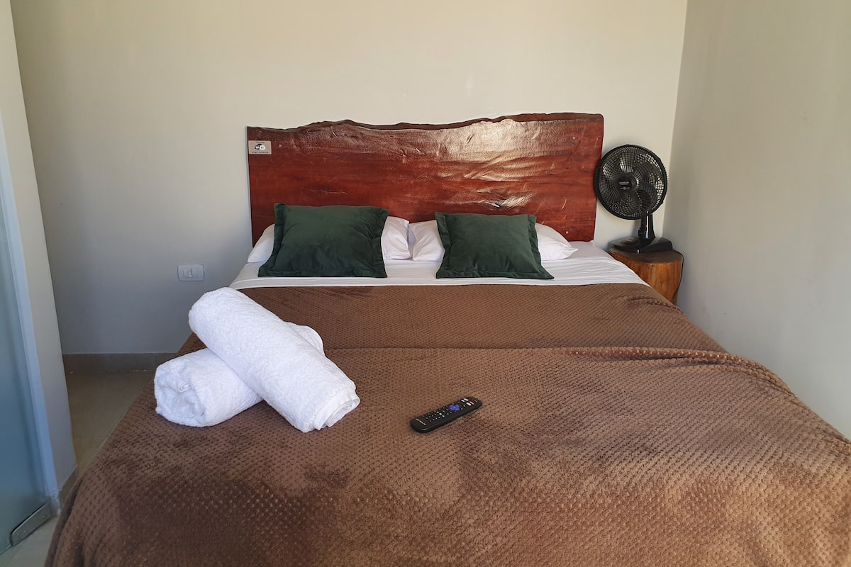 A neatly made bed with a brown blanket, two green pillows, and a white towel. A remote control is on the bed.