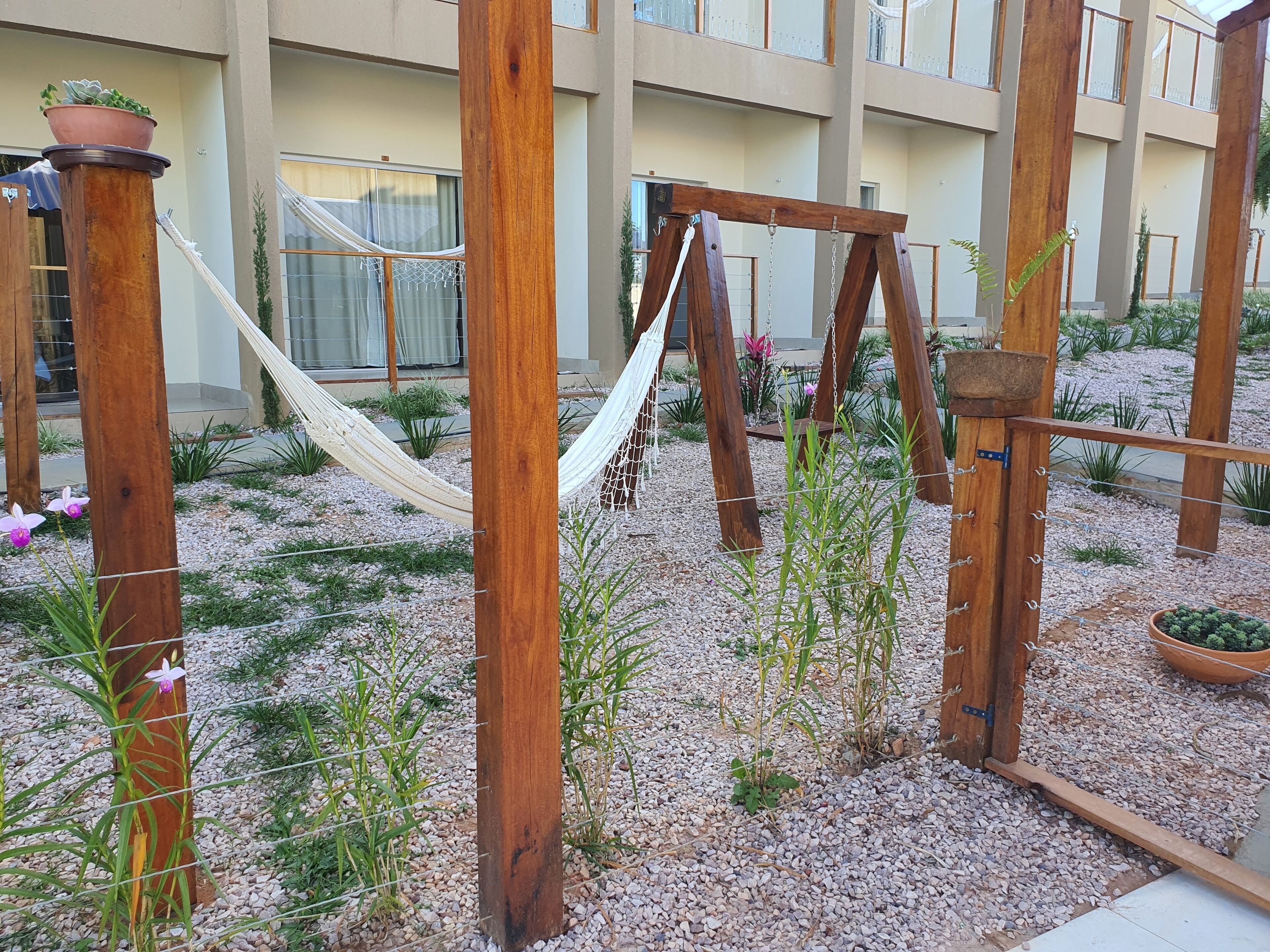 A wooden swing set in a courtyard with a hammock, surrounded by gravel and plants.