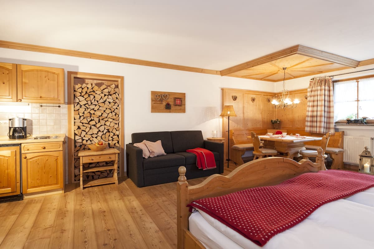 A cozy room with a bed, a wooden dresser, a small table, a sofa, and a kitchen area with a coffee maker.
