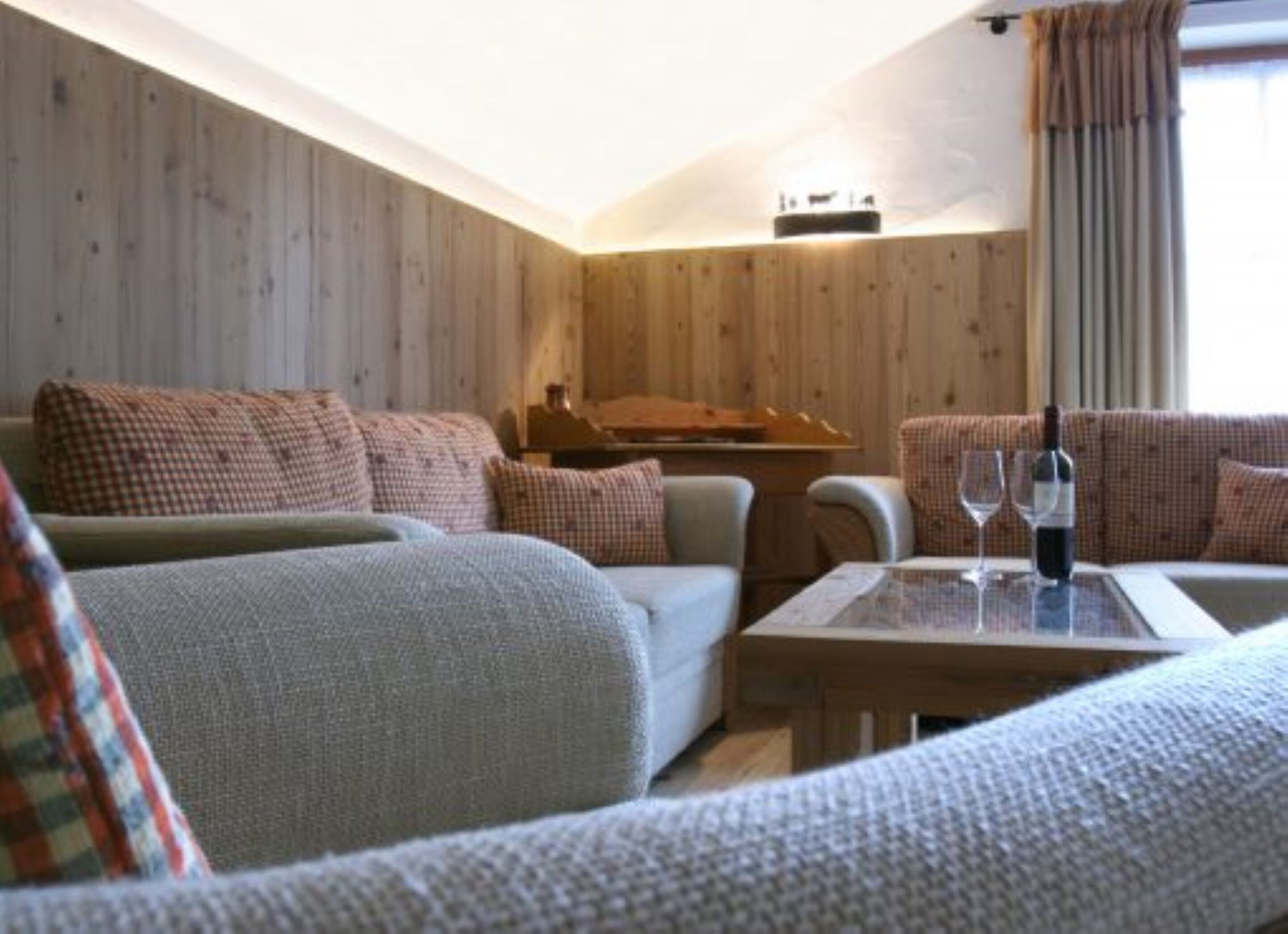 A cozy living room with a wooden wall, a sofa set, a coffee table with a bottle and glasses, and a window with curtains.
