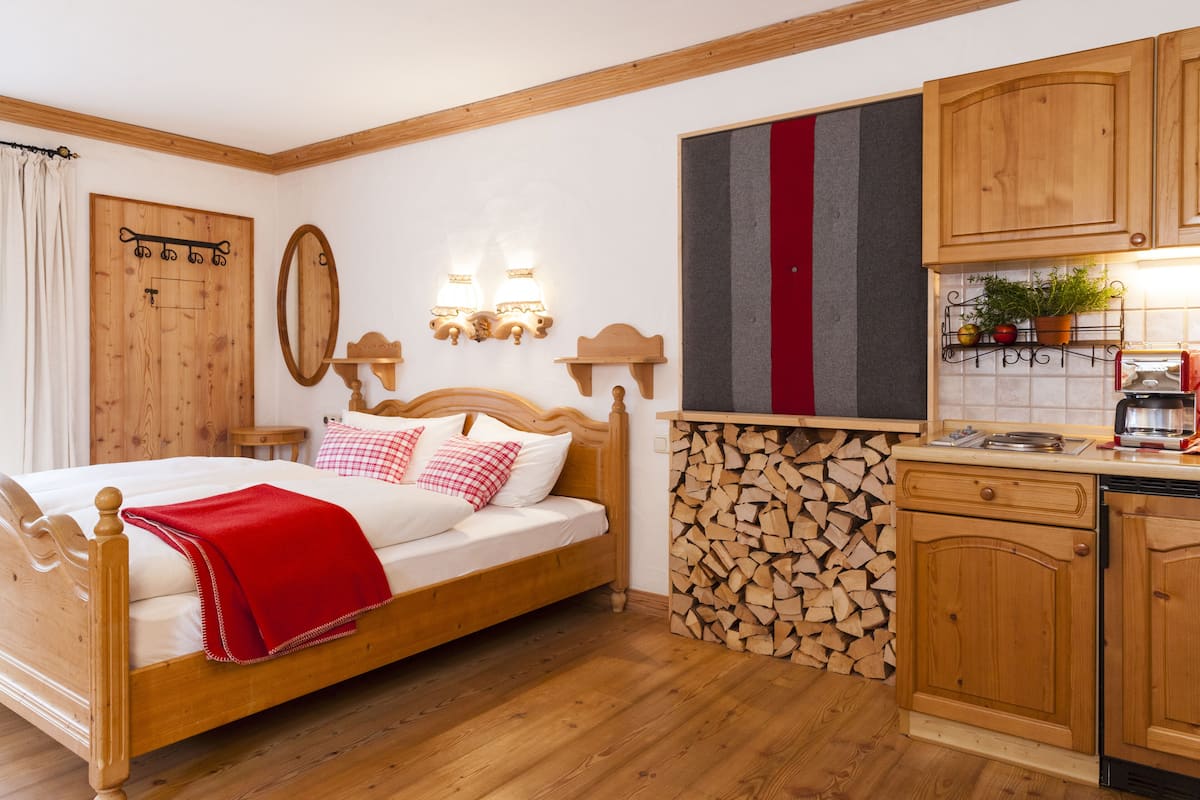 A wooden bed with a red blanket, a wooden headboard, and a bedside table with a lamp. There is a wall-mounted mirror and a wooden cabinet with a red stripe.