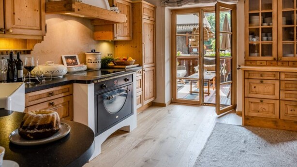 A kitchen with wooden cabinets, a central island with a stove, and a dining area visible through glass doors. - Fiakerhof (Garmisch-Partenkirchen)