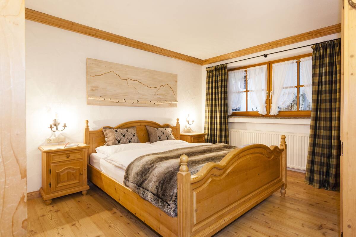 A wooden bed with a headboard, a bedside table with a lamp, a wall-mounted artwork, and a window with curtains.