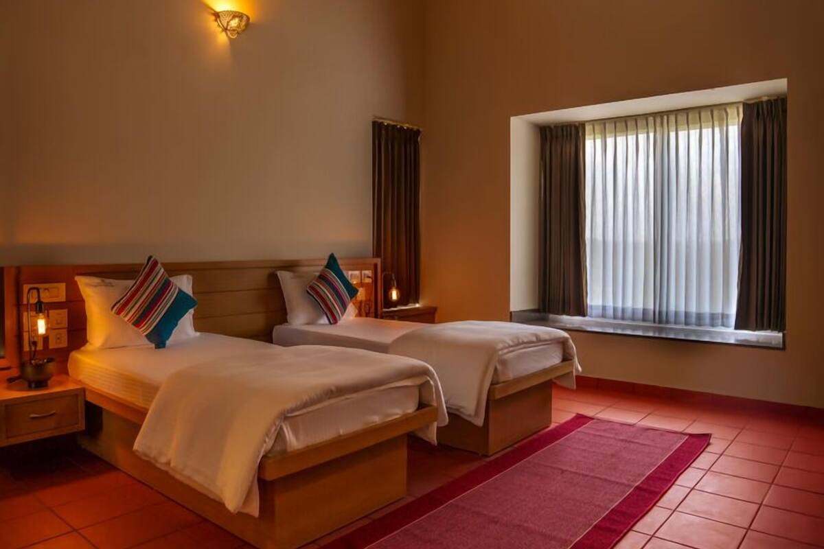 A hotel room with two beds, a red rug, a window with curtains, and a wall-mounted light fixture.