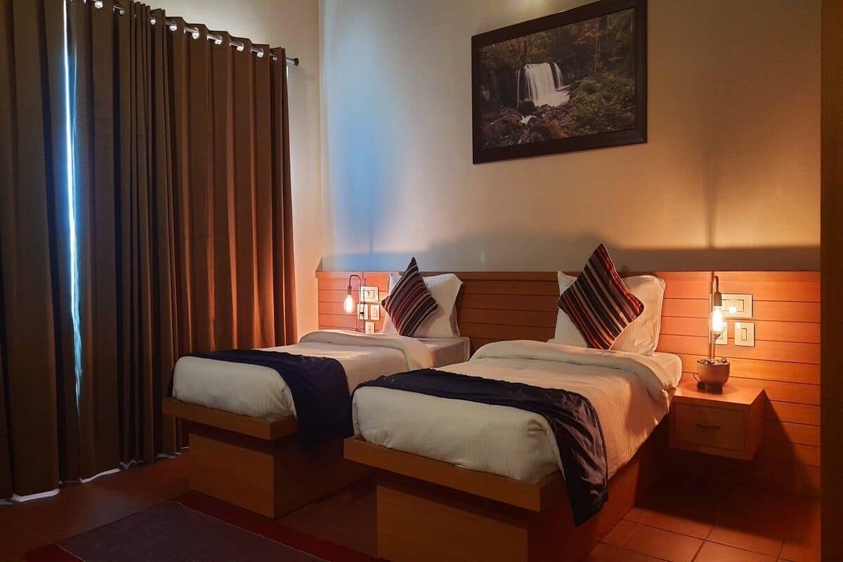 A hotel room with two beds, a nightstand with a lamp, a painting on the wall, and a window with curtains.