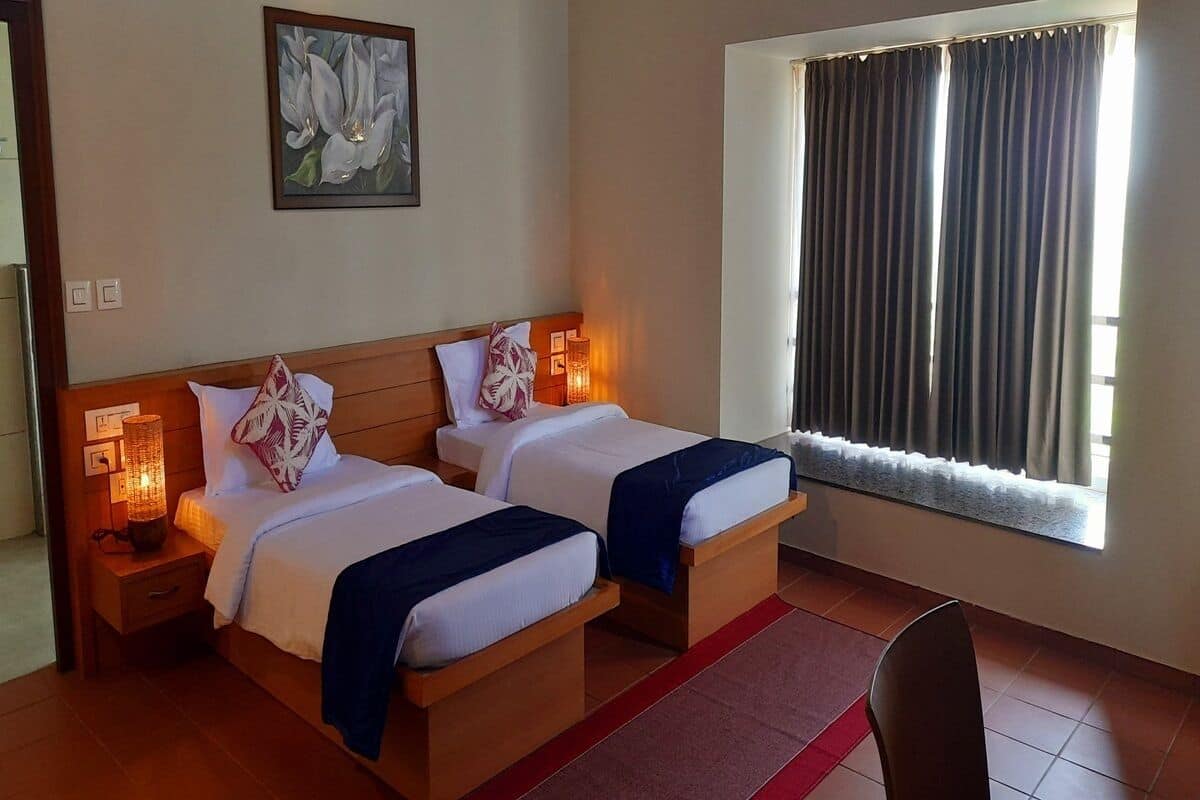 A hotel room with two beds, a nightstand with a lamp, a painting on the wall, and a window with curtains.