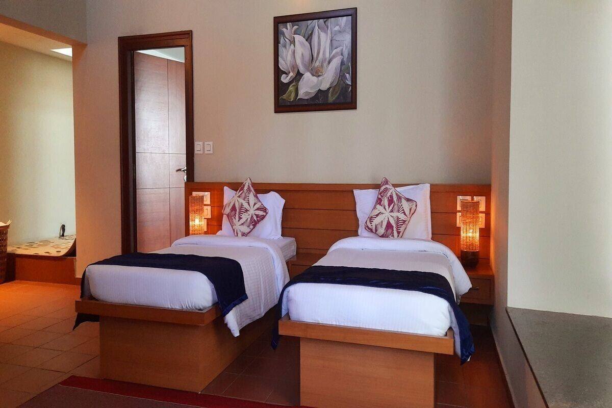 A double room with two beds, a wooden headboard, a painting of flowers, and a nightstand with a lamp.