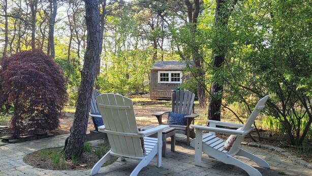 A patio area with Adirondack chairs and a small table, surrounded by trees and a wooden structure in the background.