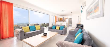 A modern living room with a large window, grey sofas with colorful cushions, a wooden coffee table, and a view of the city.