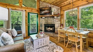 Interior acogedor de una cabaña con chimenea de piedra, muebles de madera y ventanas amplias con vistas al bosque.