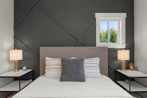 A modern hotel room with a large bed, two bedside tables with lamps, a window with a view of trees, and a geometric wall design.