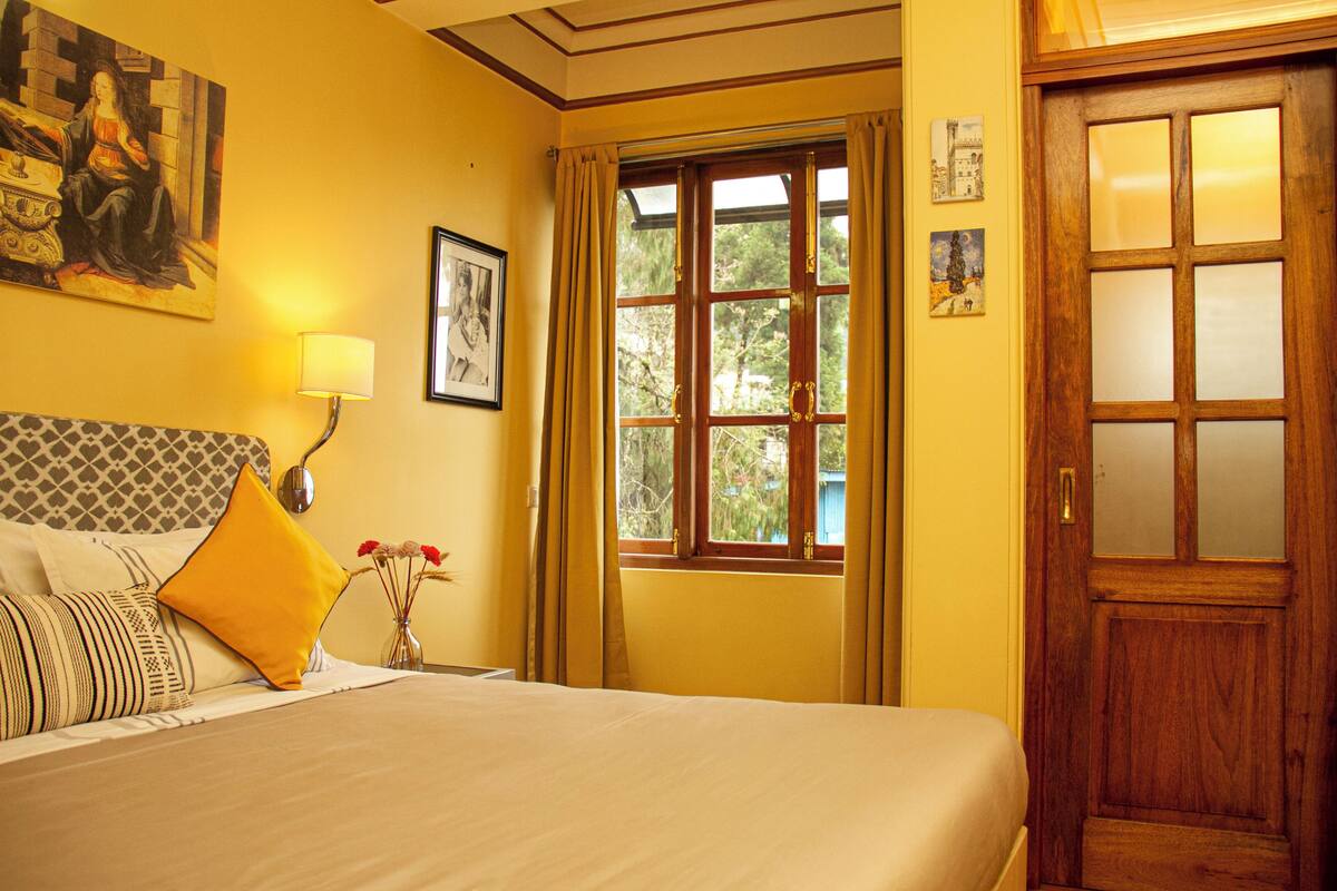 A hotel room with a bed, a nightstand, a lamp, a window with curtains, and a wooden door.