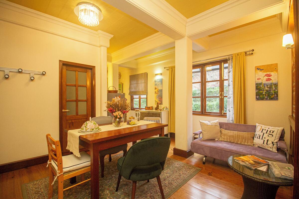 A dining area with a wooden table, chairs, and a vase of flowers. There is a sofa and a small table with magazines.