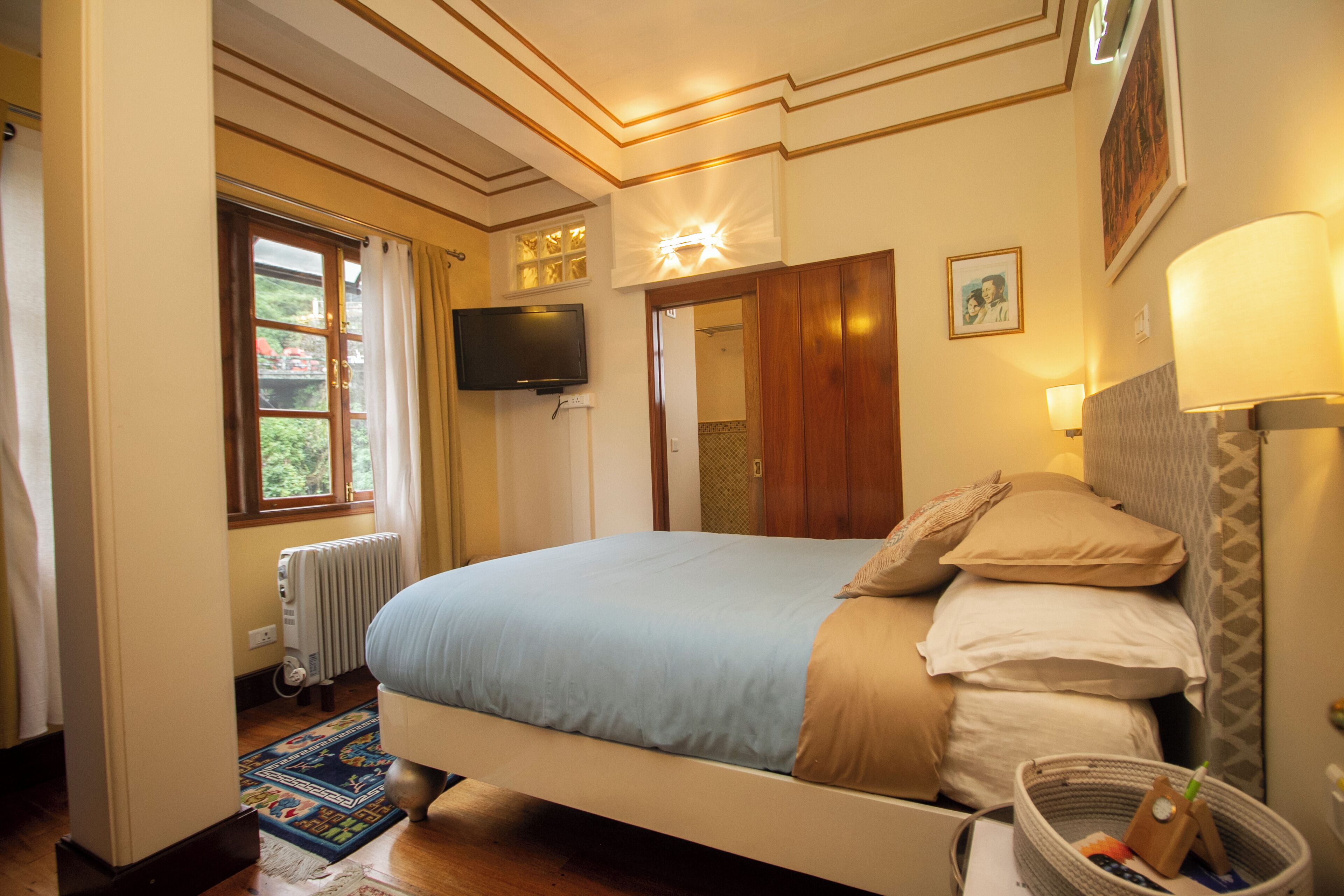A bedroom with a bed, a television, a window with curtains, a radiator, and a bedside lamp.