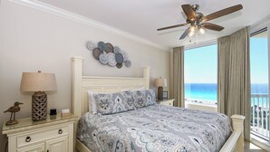 Een slaapkamer met een bed, twee nachtkastjes, een plafondventilator, een decoratief wandobject en uitzicht op de oceaan.