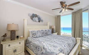 A bedroom with a bed, two bedside tables, a ceiling fan, a decorative wall piece, and a view of the ocean. - Beautiful Gulf Views from 4th Floor of Silver Shells St. Croix! (Destin)