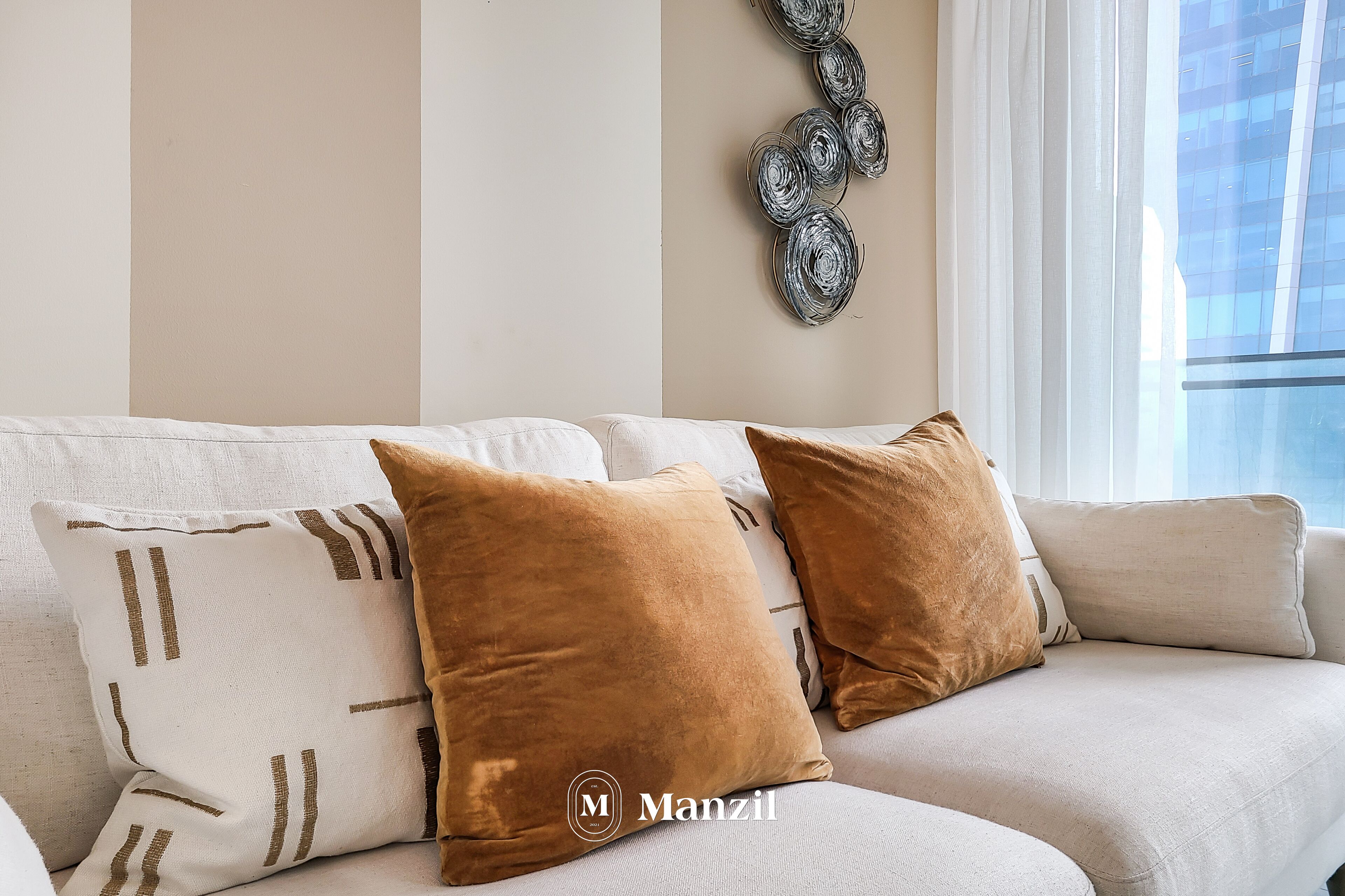 Ein modernes Wohnzimmer mit einem beigen Sofa, zwei braunen Kissen und einer dekorativen Metallwandarbeit.