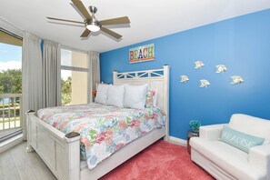 Um quarto com uma cama, um ventilador de teto, uma janela com cortinas, uma parede com a placa 'BEACH' e um sofá branco com uma almofada 'PARADISE'.