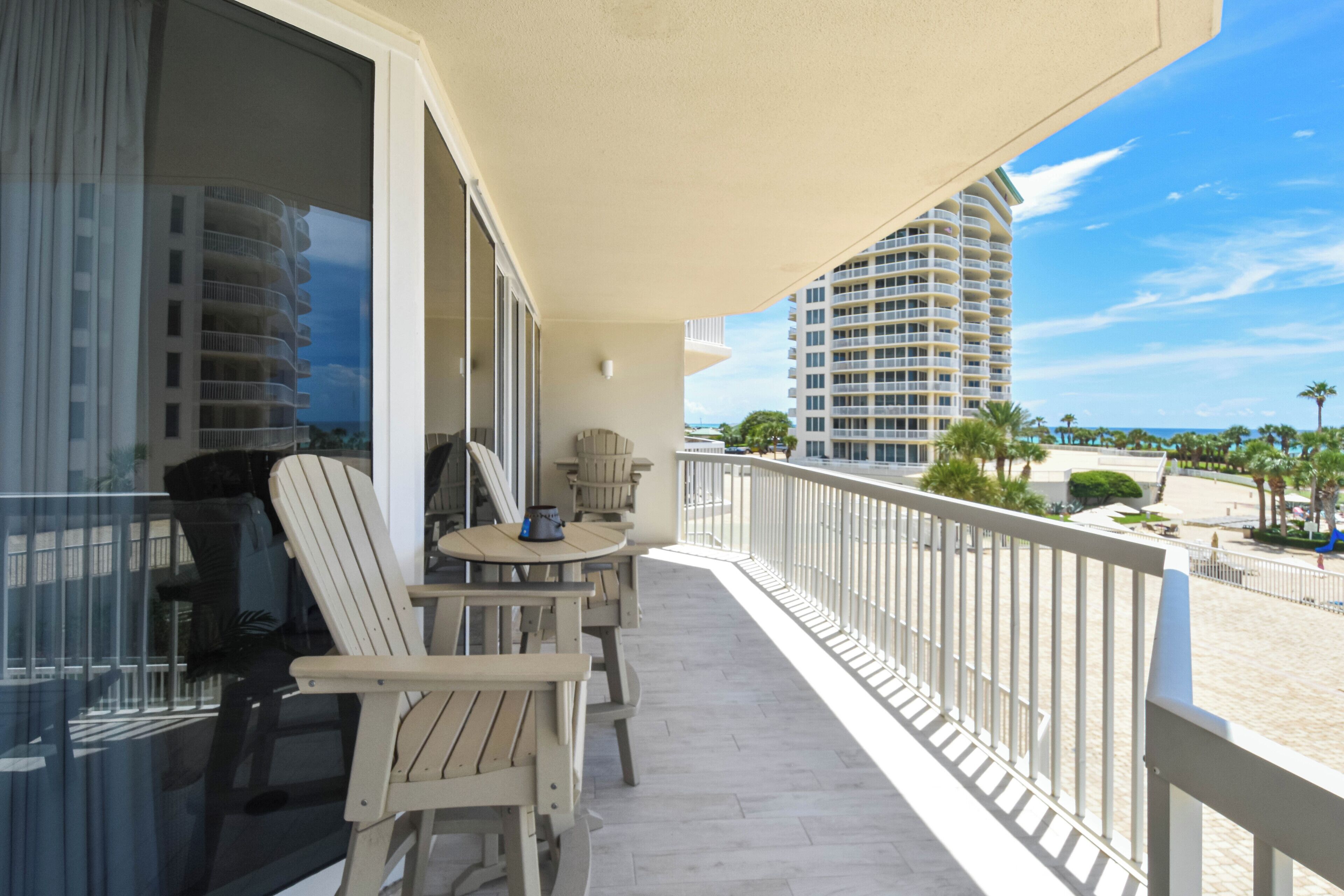 A balcony with two Adirondack chairs, a small round table, and a view of a tall building and beach.