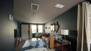 A living room with a sofa, coffee table, and lamp. There are two windows with curtains, a door, and a wall-mounted mirror. - Haus of Style Moody Blues (Charlotte)