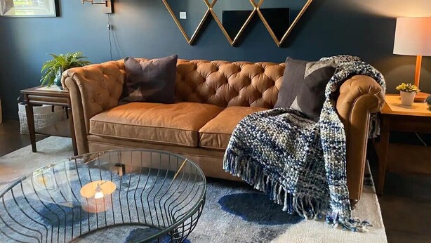 A modern living room with a brown leather sofa, a glass coffee table, and a geometric wall mirror. - Haus of Style Moody Blues (Charlotte)